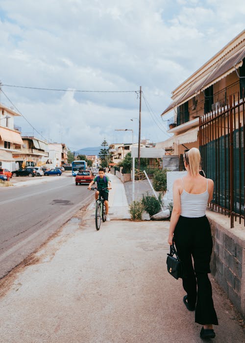 Back View of a Woman Walking on a Sidewalk and a Boy Riding a Bicycle in a Town 