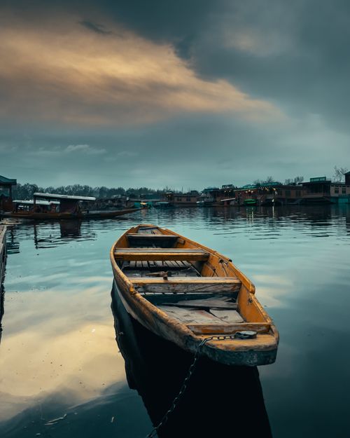 Free A wooden boat moored on a calm lake under an overcast sky, reflecting a serene village setting. Stock Photo