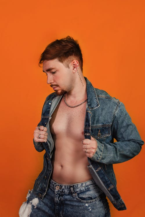 Shirtless Man Wearing Jeans and Denim Jacket Posing on Orange Background in Studio 