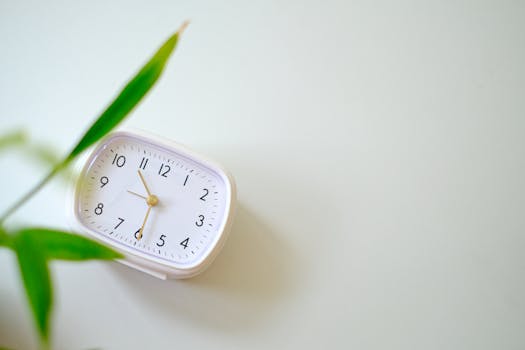 A minimalist white desk clock paired with a green leaf on a soft background.