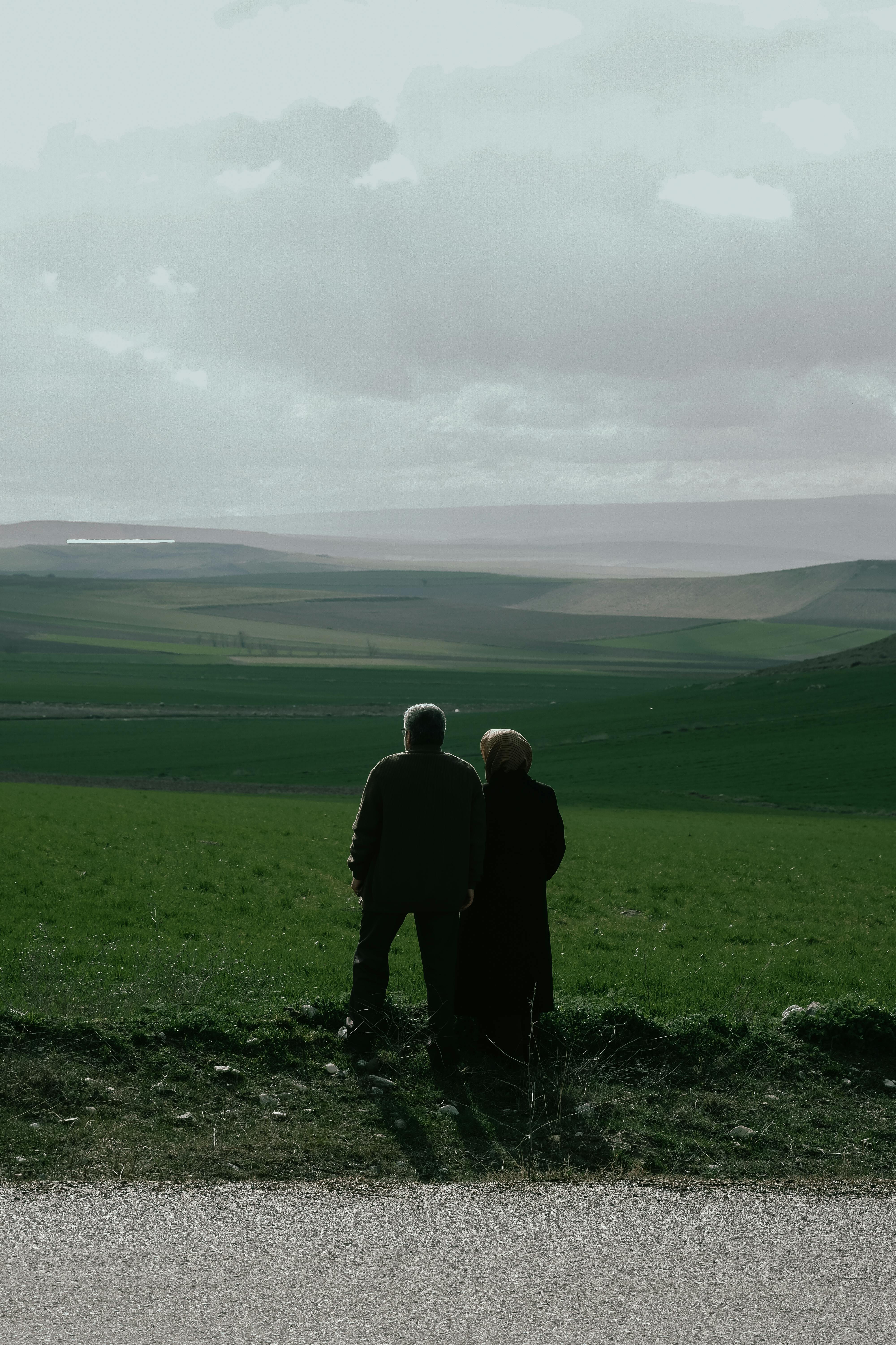 A man and woman stand overlooking a wide green valley under a cloudy sky.