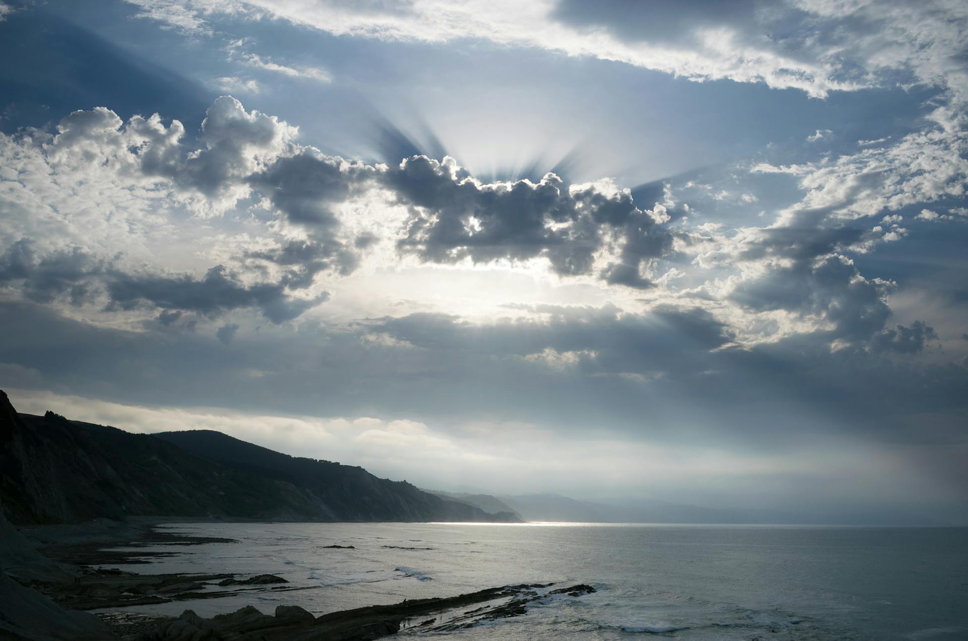 Scenic view of ocean and coast under dramatic cloud-filled sky with sun rays.