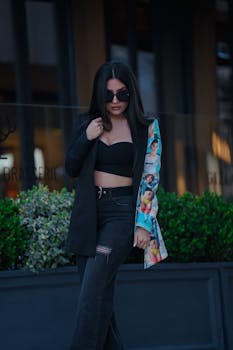 Trendy woman in black top and blazer posing outdoors with sunglasses.