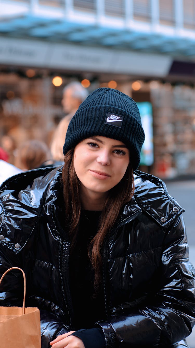 Young Woman In A Winter Jacket And Hat Sitting Outside In City 