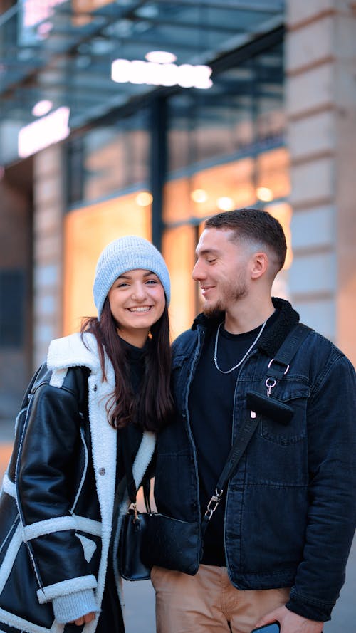 A Young Couple in Warm Clothing Standing in front of a Building in City and Smiling 