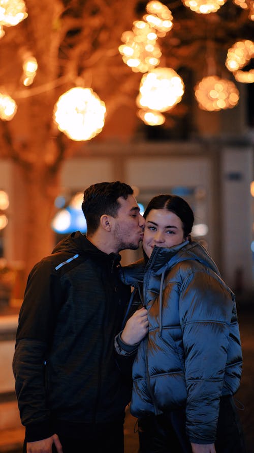 Man Kissing a Woman on the Cheek on the Background of Lights in City in the Evening 