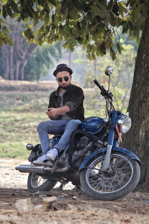 Free A man in sunglasses and hat sitting on a motorcycle under a tree using his phone. Stock Photo