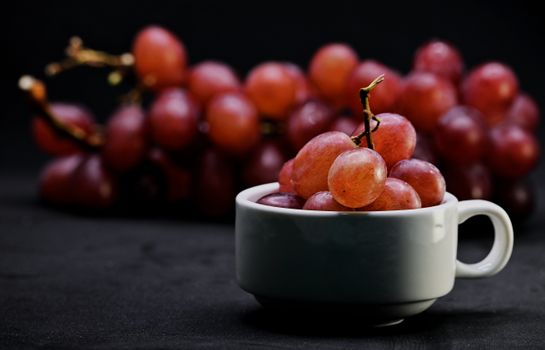 A close-up image of red grapes piled in a white cup with more grapes blurred in the background, set against a dark backdrop.