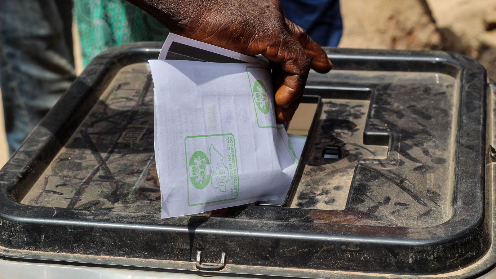 A close-up shot of a hand placing a paper ballot into a transparent voting box outdoors.