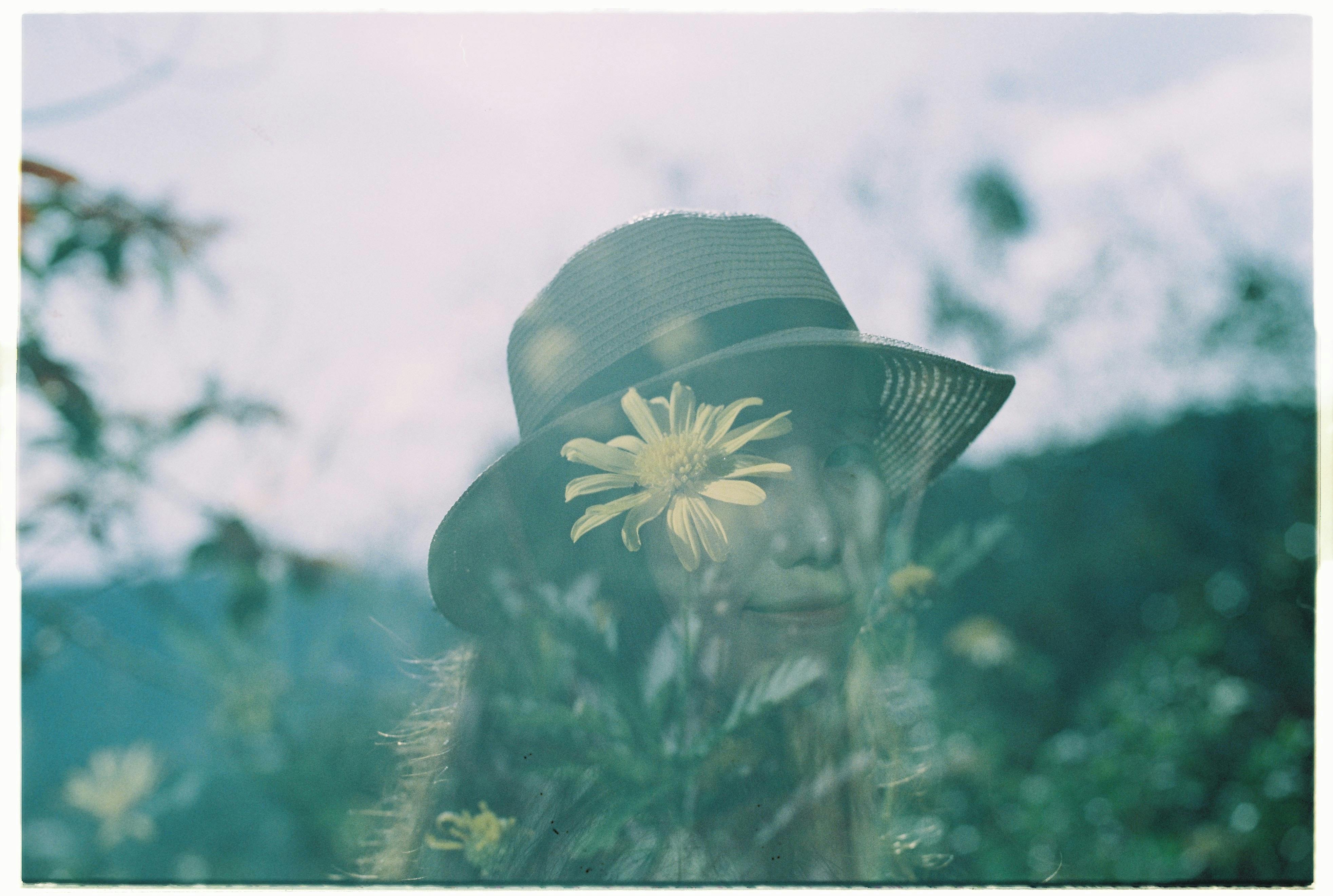 A double exposure of a woman in a straw hat with a yellow flower overlay outdoors.