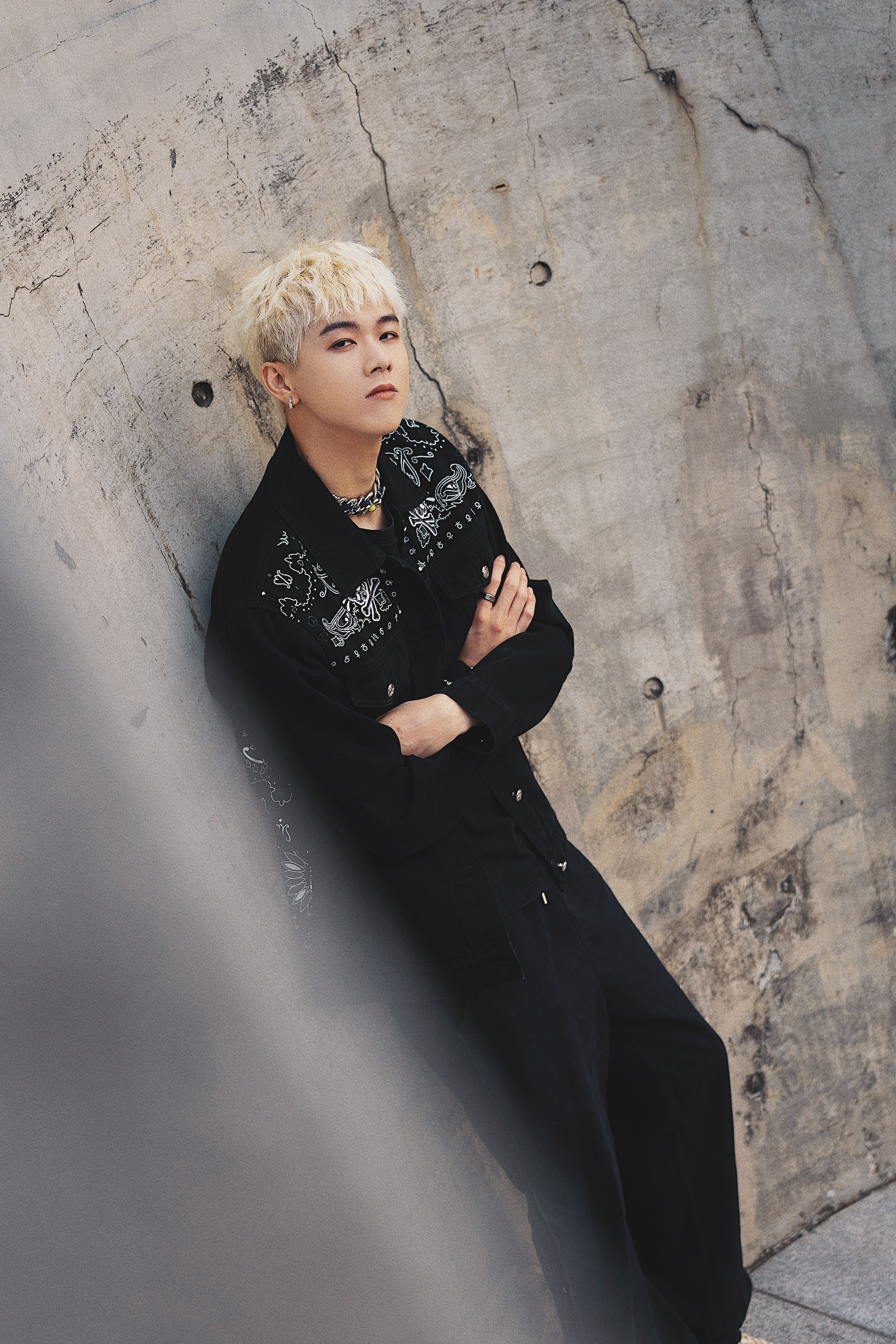 Blonde person in stylish black outfit leaning on urban concrete wall, exuding a trendy fashion vibe.
