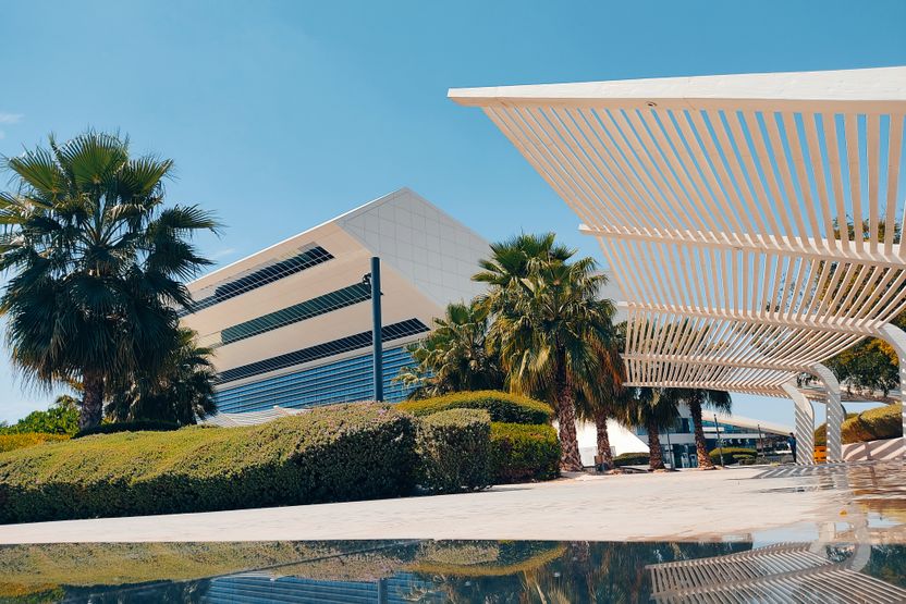 A modern resort building under a deep blue sky, with a wide white louvred canopy, a long low white block behind, mature palms between them and a still reflecting pool in front.