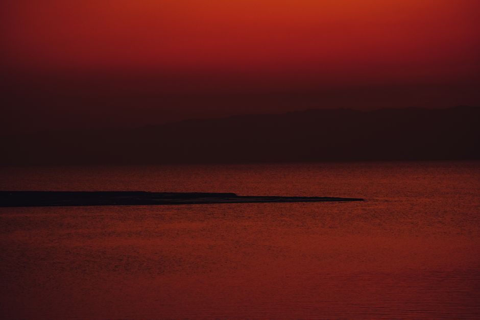 A striking red sunset creates a dramatic, tranquil seascape with a distant peninsula.