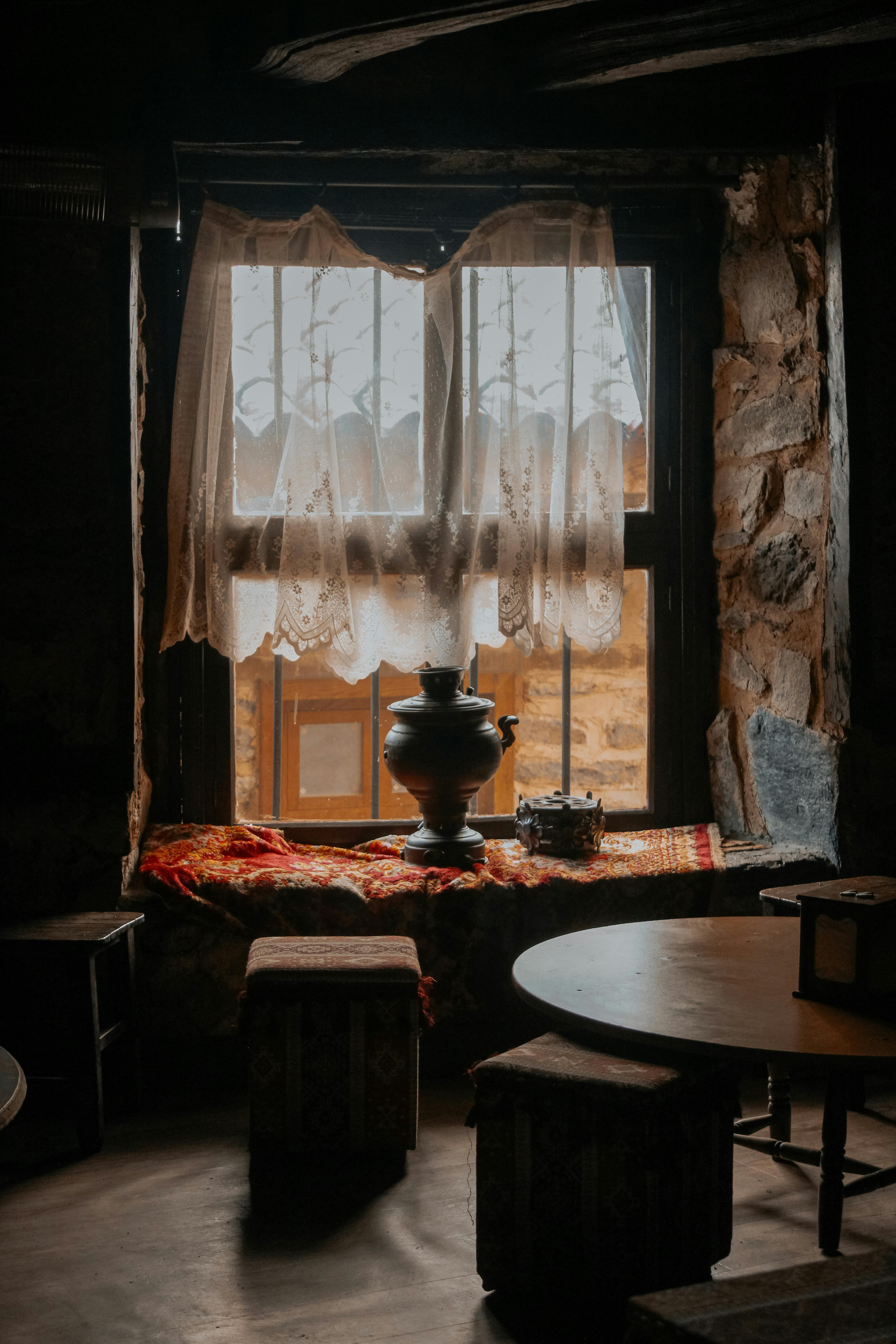 A warm, rustic room features lace curtains and a samovar by the window, creating a cozy atmosphere.