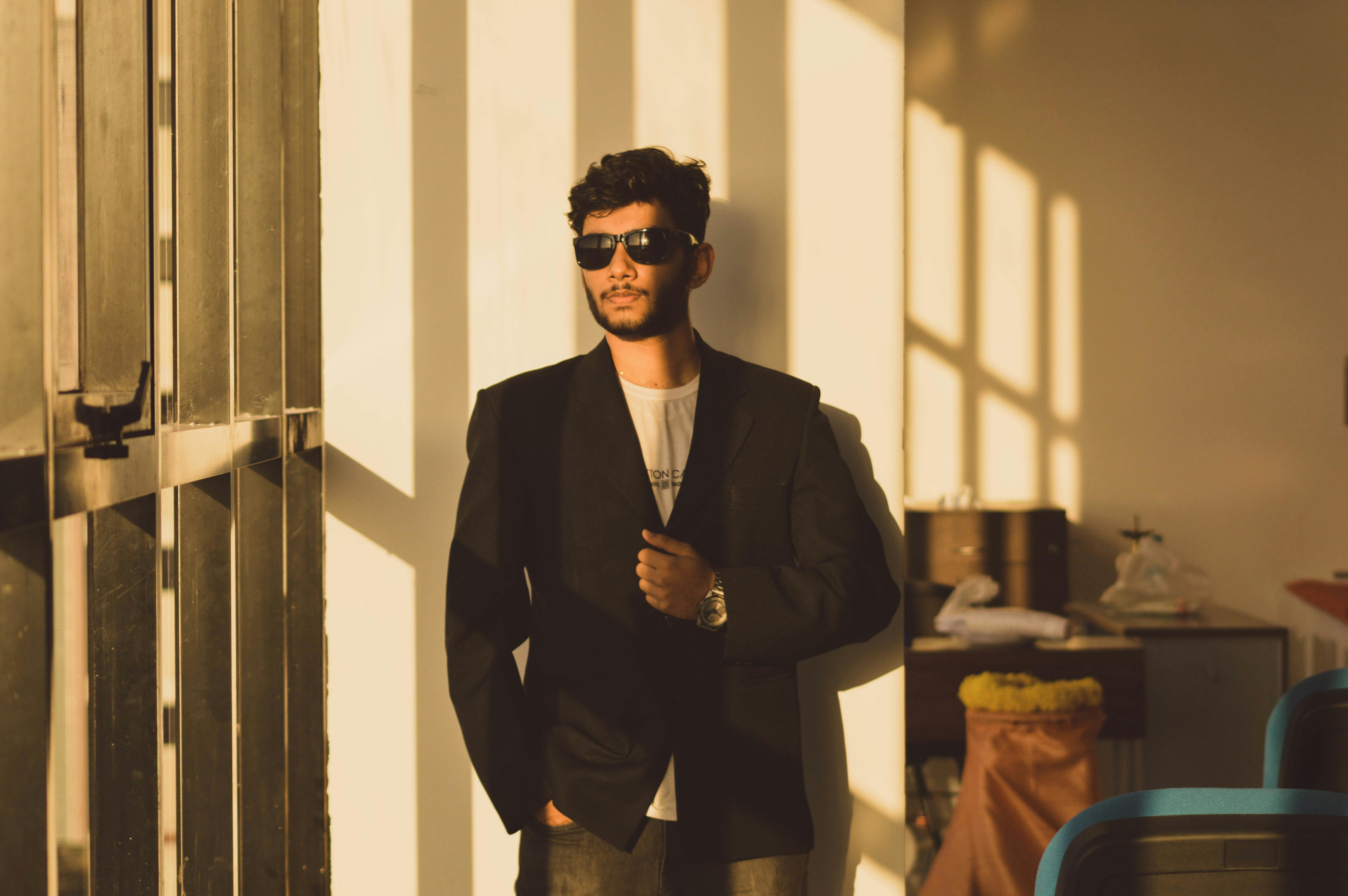 一位戴着墨镜、穿着西装的时尚男士自信地倚在阳光照射的墙上