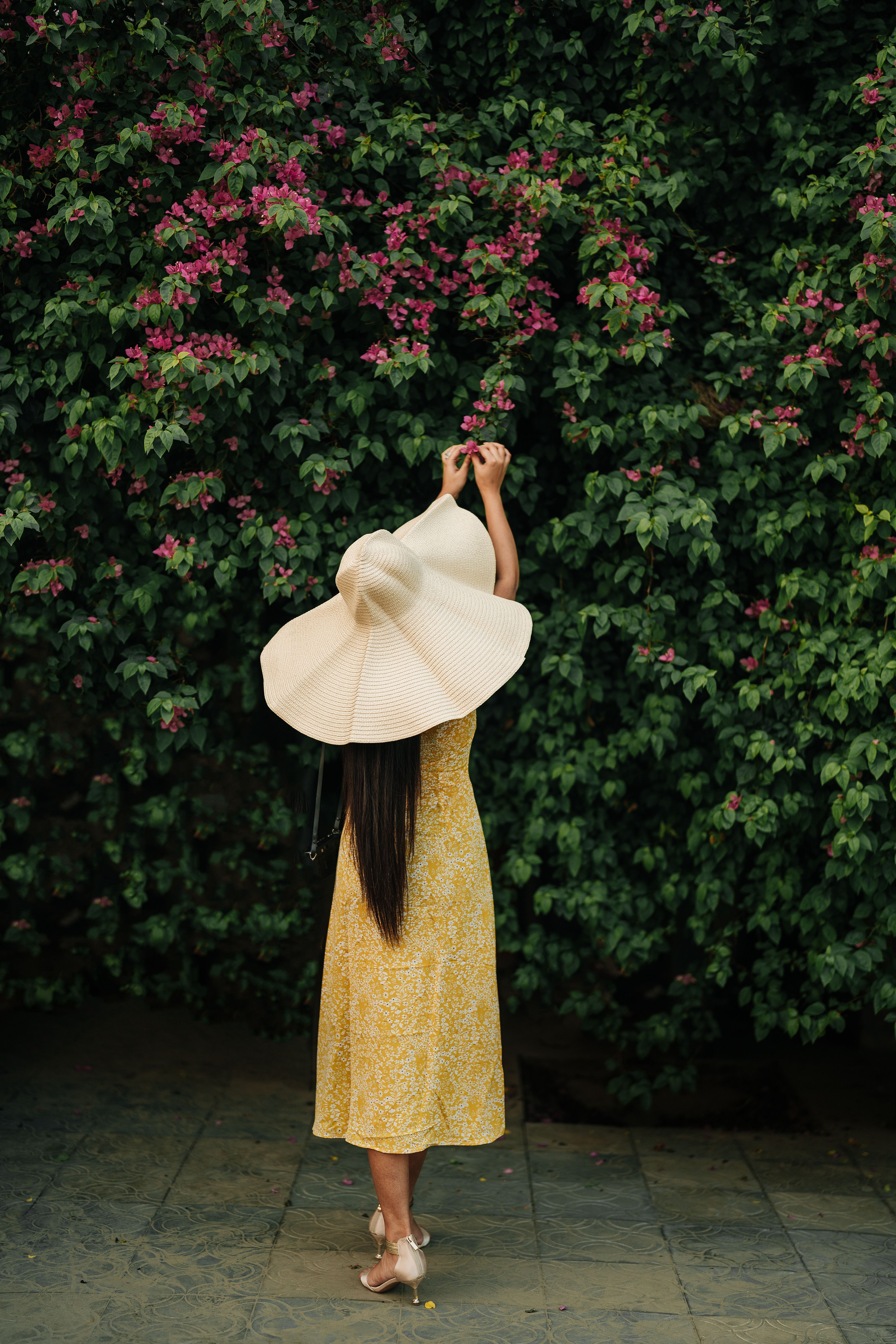 A woman in a yellow dress with a sun hat reaching for pink flowers against a lush green shrub.