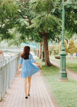 一位身着蓝色连衣裙的女子，正沿着绿树环绕的公园小径行走，这是她的背影