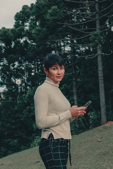Fashion portrait of a woman standing with smartphone in a forest setting.