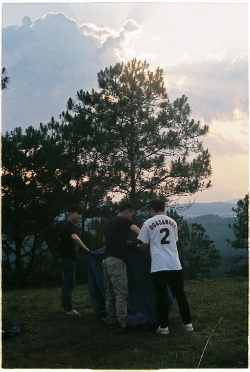 Free A group of adults is setting up a tent in a grassy area surrounded by trees and a cloudy sky. Stock Photo