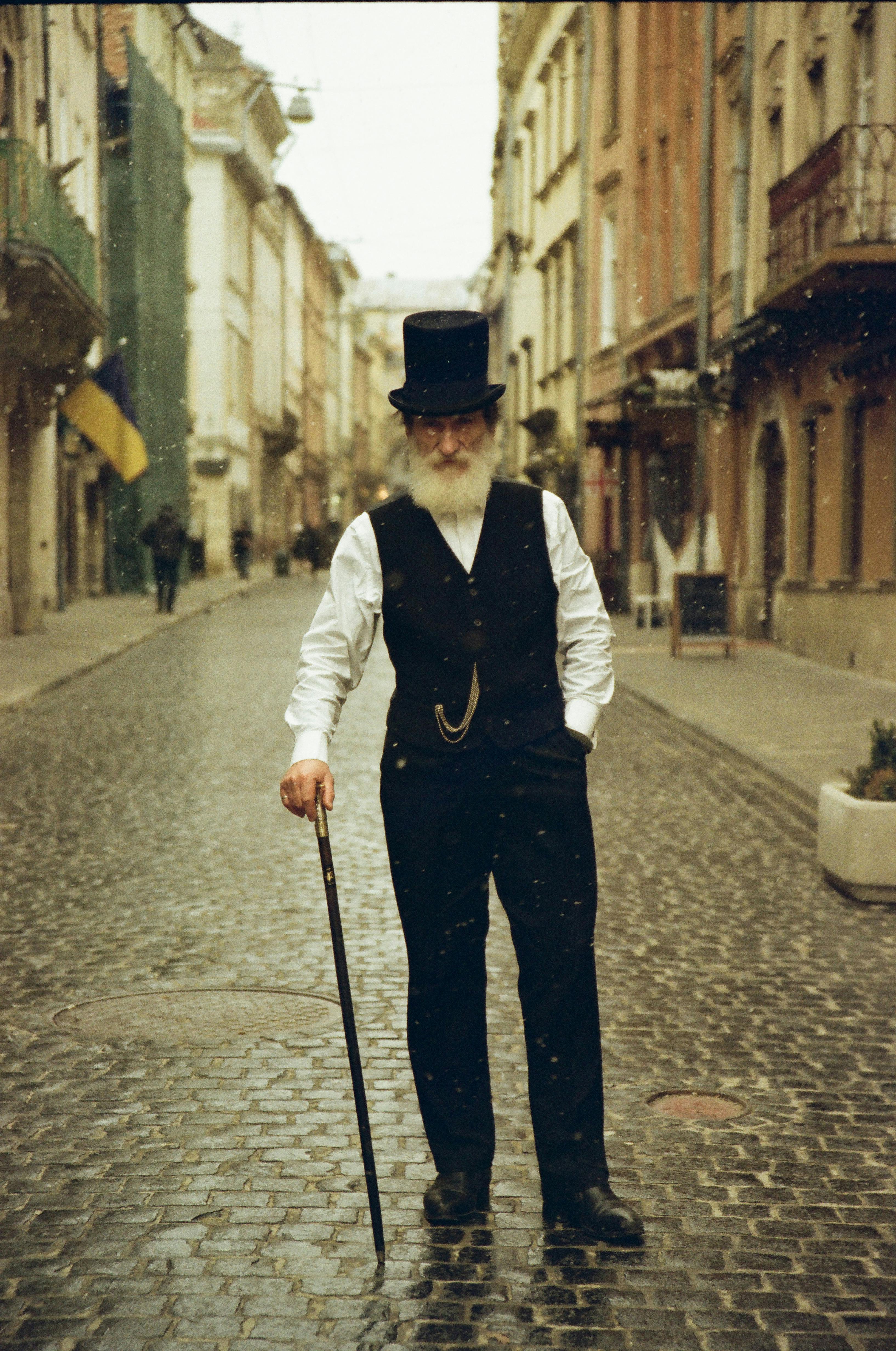 A distinguished elderly man with a cane walks in a historic European street dressed elegantly in vintage attire.