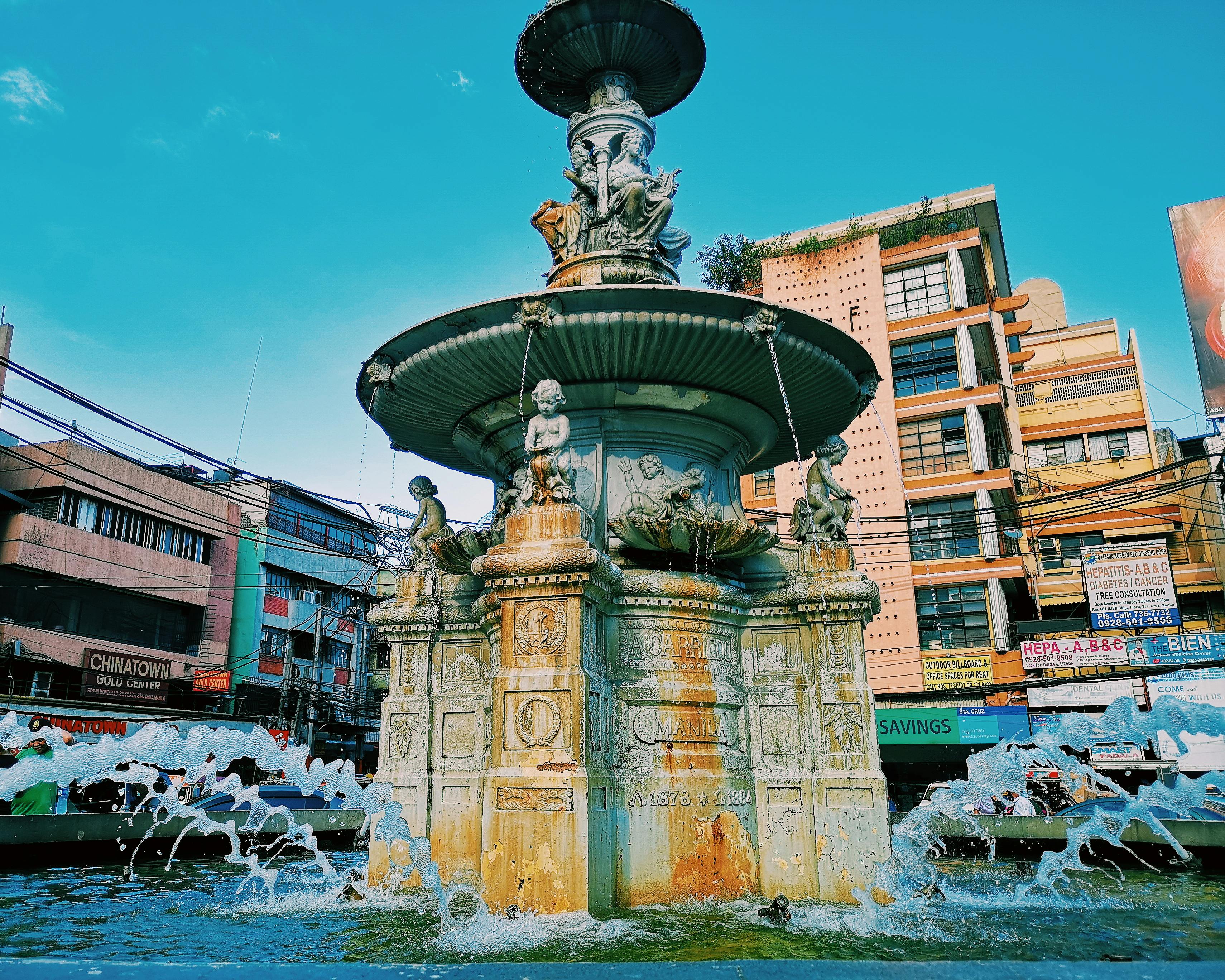 城市建筑环绕中，一座宏伟的雕塑喷泉矗立在晴朗的蓝天下