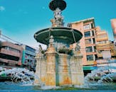 城市建筑环绕中，一座宏伟的雕塑喷泉矗立在晴朗的蓝天下