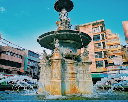 城市建筑环绕中，一座宏伟的雕塑喷泉矗立在晴朗的蓝天下