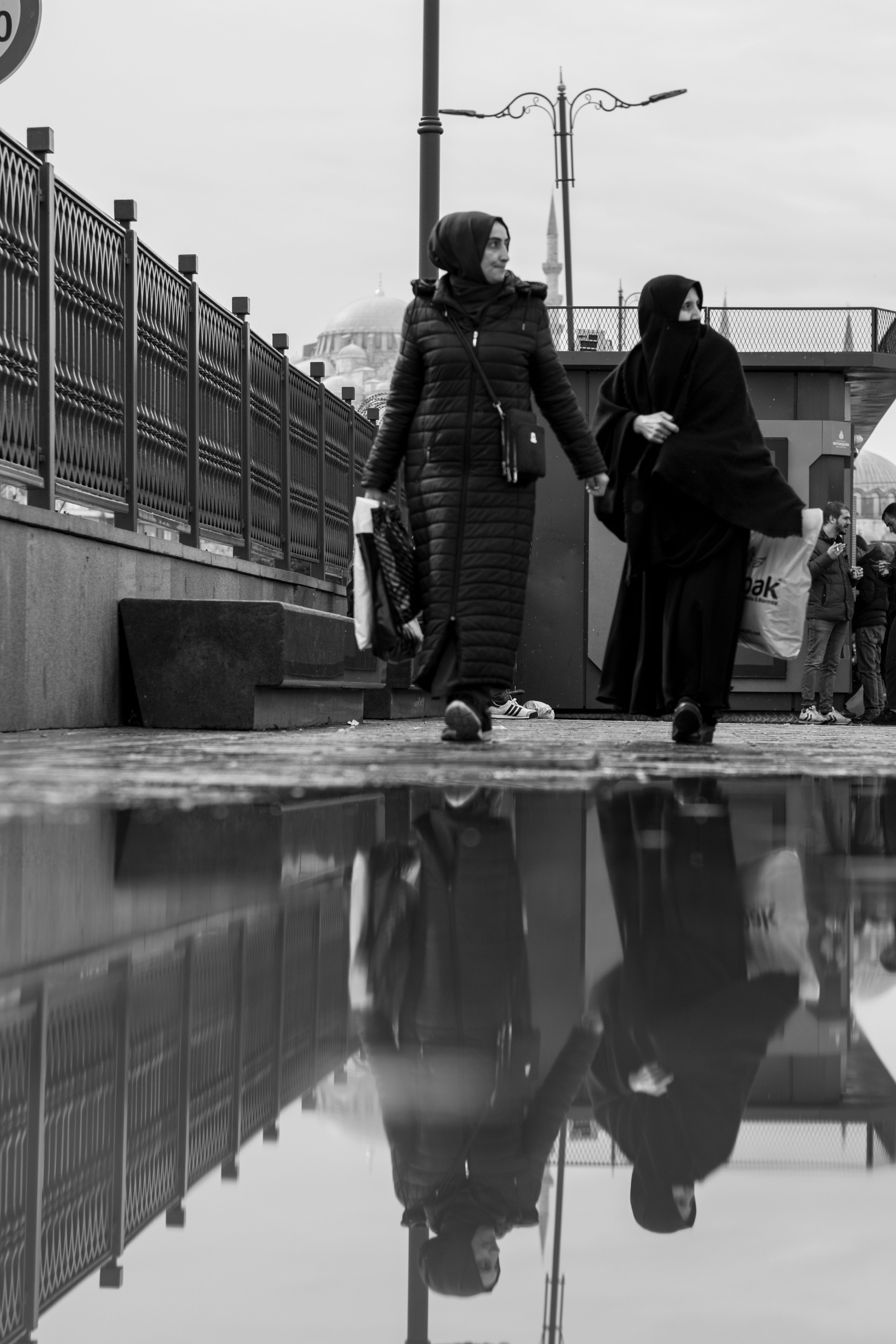 Black and white photo of two women walking with reflections in a puddle.