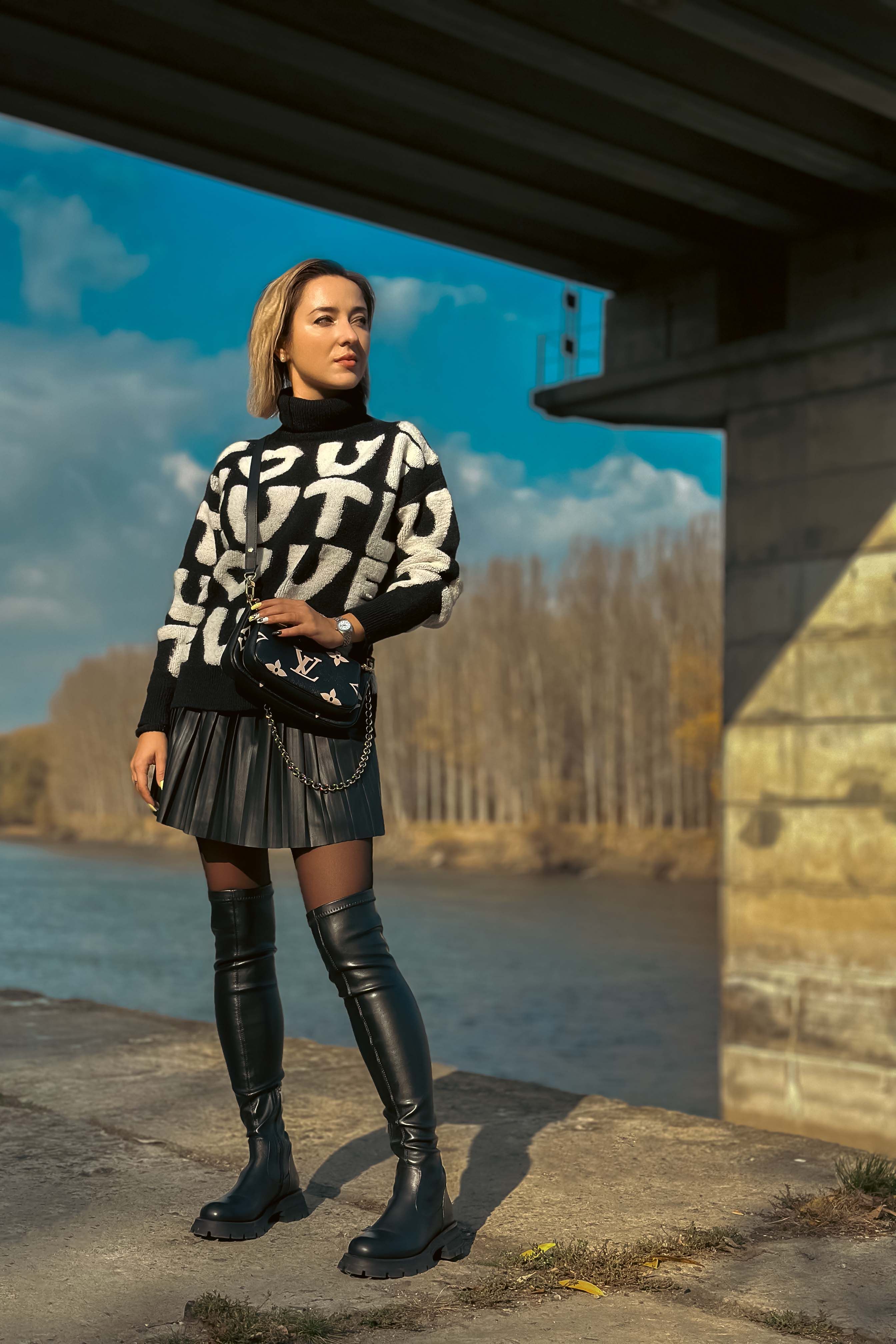 Woman in sweater and boots stands under a bridge by the river on a sunny day.