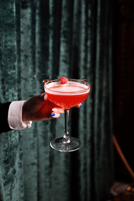 A round black Chambord bottle with its gold crown-shaped neck label next to a pink cocktail garnished with fresh raspberries
