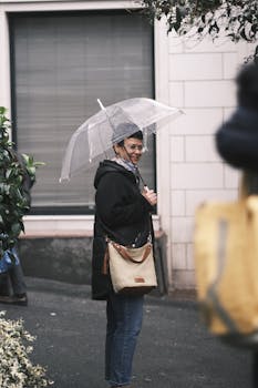 A woman smiles while standing on a city sidewalk holding a clear umbrella on a rainy day.