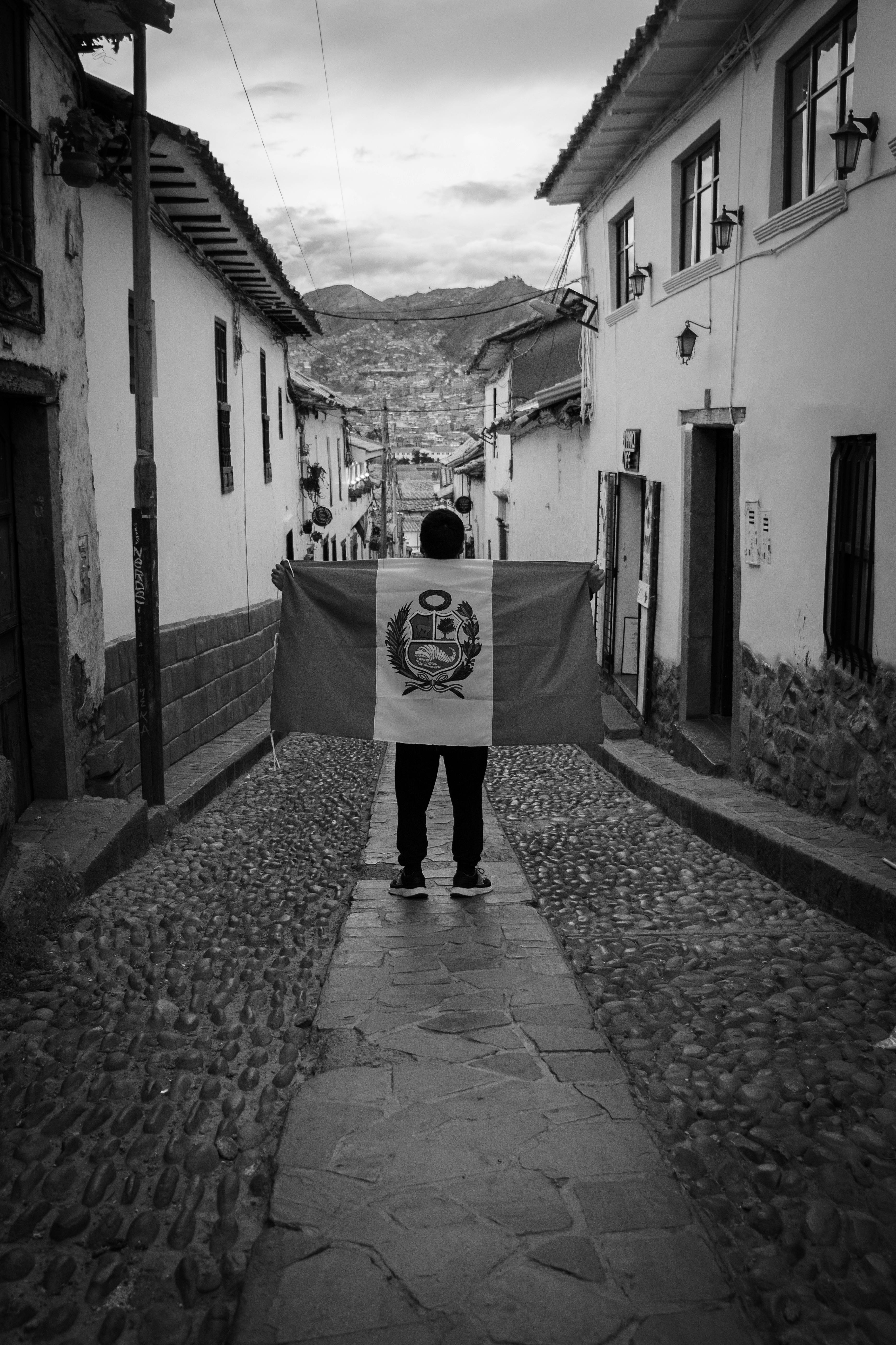 Black and white photo of a person holding the Peruvian flag on a cobblestone street in Cusco.