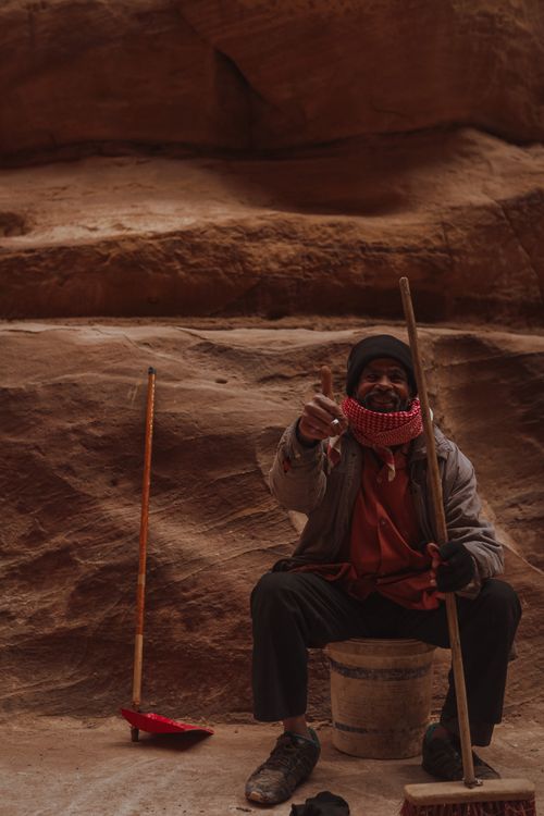 Free Elderly man sitting on a bucket in Petra, Jordan, with traditional attire and broom, giving a thumbs up. Stock Photo