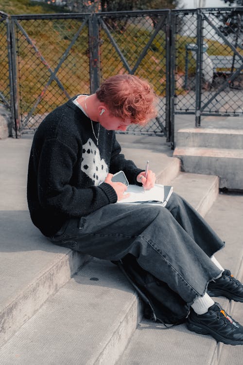 A Boy Sitting on Steps and Writing in the Notebook 