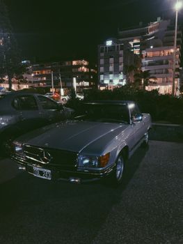Classic silver Mercedes Benz R107 parked at night with city buildings in background.