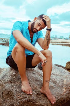 Man with tattoos sitting barefoot on a rock by the beach, showcasing fashion and casual style.