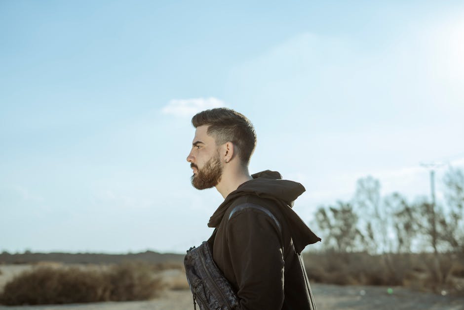 A stylish young man with a beard and mustache in an outdoor setting, dressed casually.