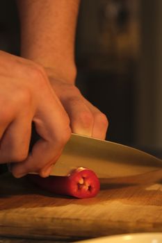 A person's hands cutting a red chili pepper on a wooden board in warm lighting.
