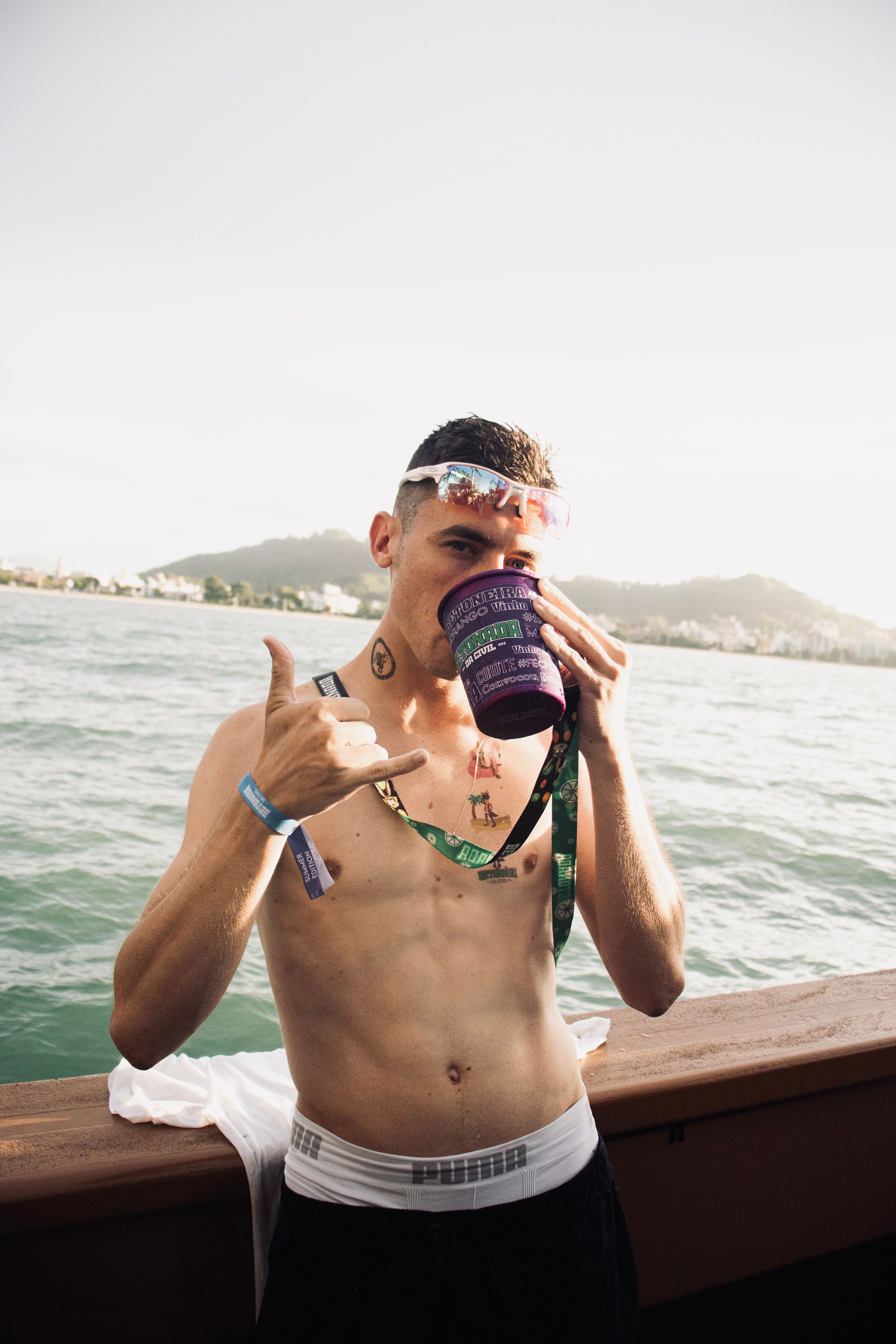 A shirtless young man in sunglasses sips from a cup on a boat under the sun.
