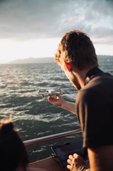 Back view of a man holding a drone over the ocean during sunset, with overcast skies.