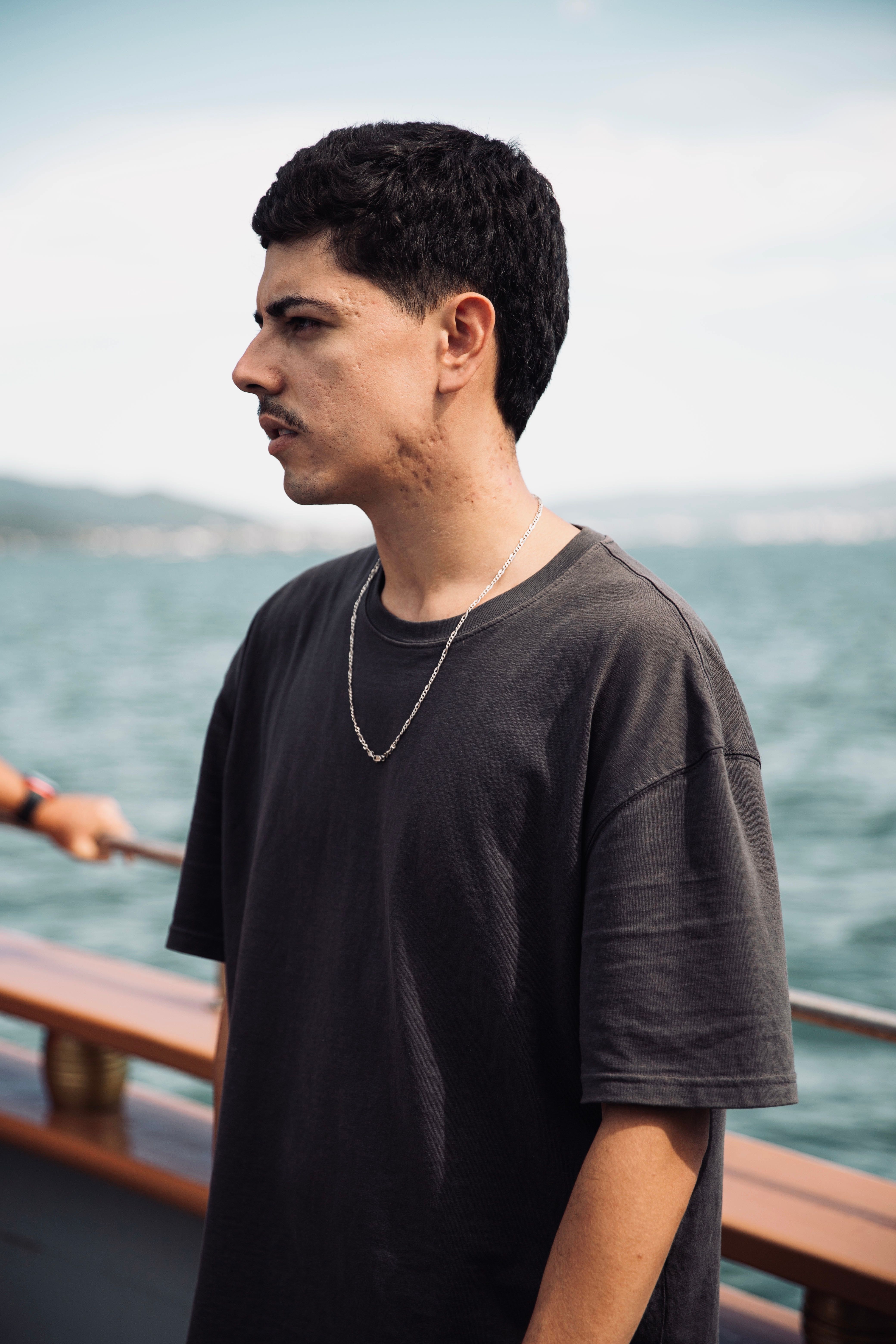 Man with short black hair wears necklace and t-shirt, standing on boat with ocean view.