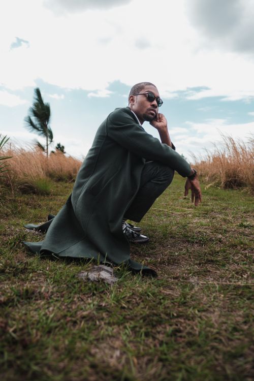 Free A stylish man in a black coat and sunglasses crouching in an open grassland, showcasing elegance and confidence. Stock Photo