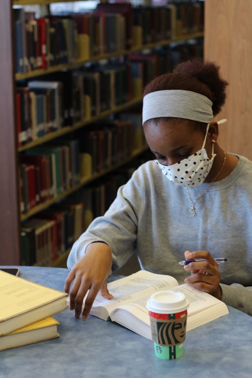 Free A young woman in a library, wearing a face mask, studying with books and a coffee. Stock Photo