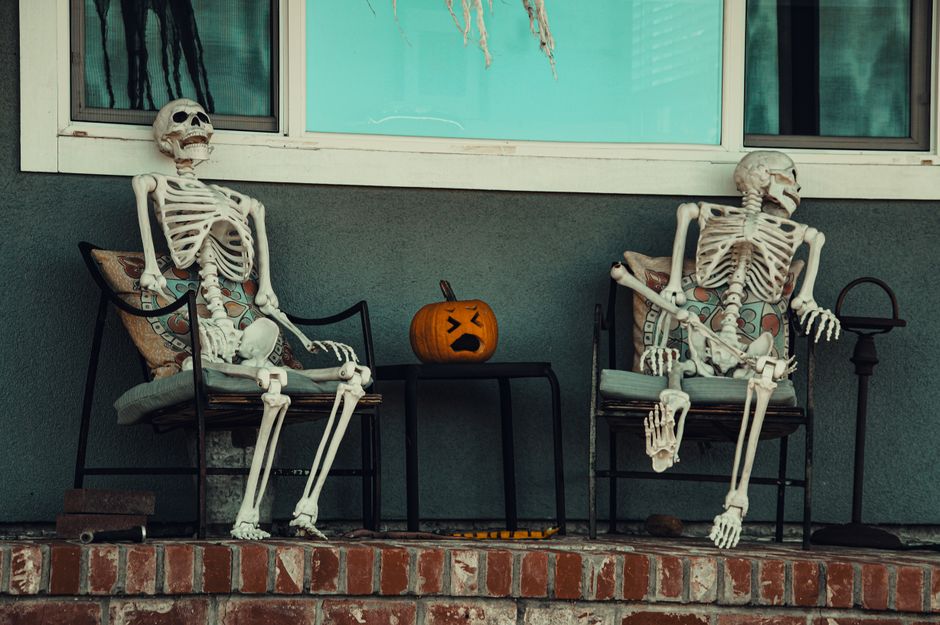 Two skeletons seated on porch chairs with a carved pumpkin, creating a Halloween atmosphere.