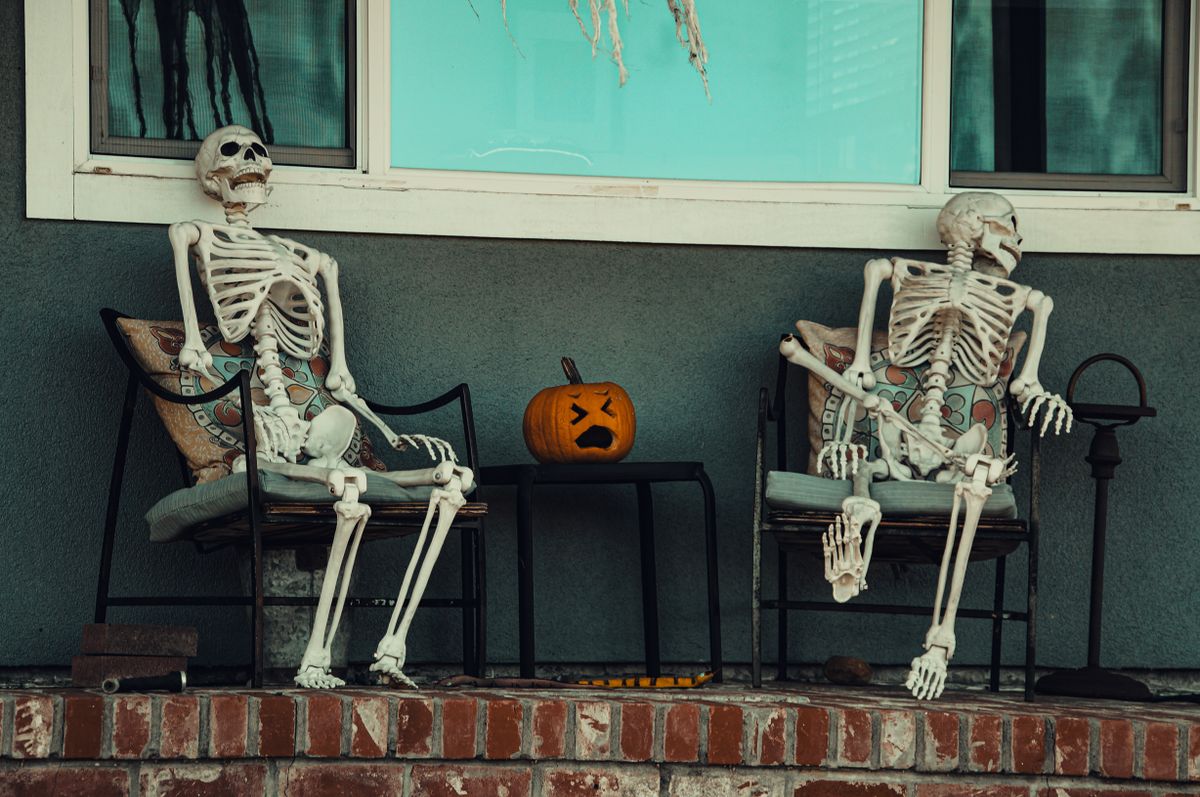 Two skeletons seated on porch chairs with a carved pumpkin