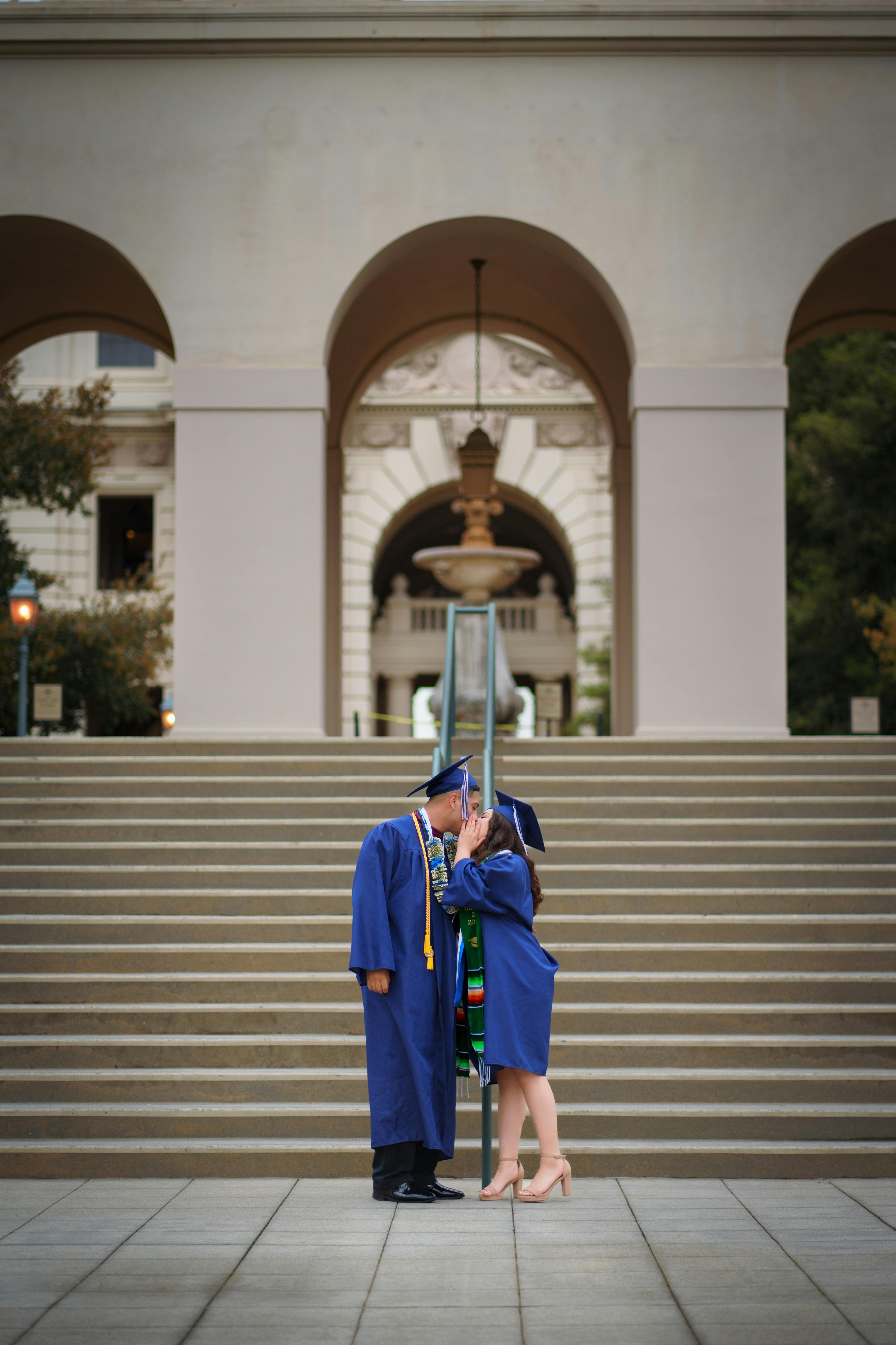 A couple of graduates in blue gowns share a tender kiss on campus steps.
