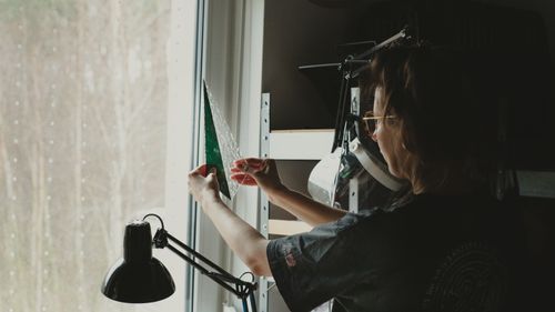Free Woman holding a triangle piece of stained glass near a window, engaging in artistic crafting. Stock Photo