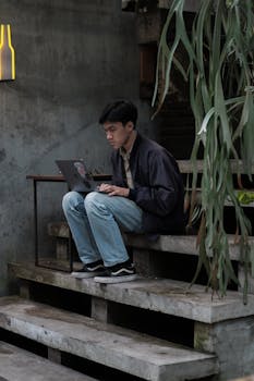 Asian man focused on his laptop while sitting on outdoor concrete steps, surrounded by greenery.
