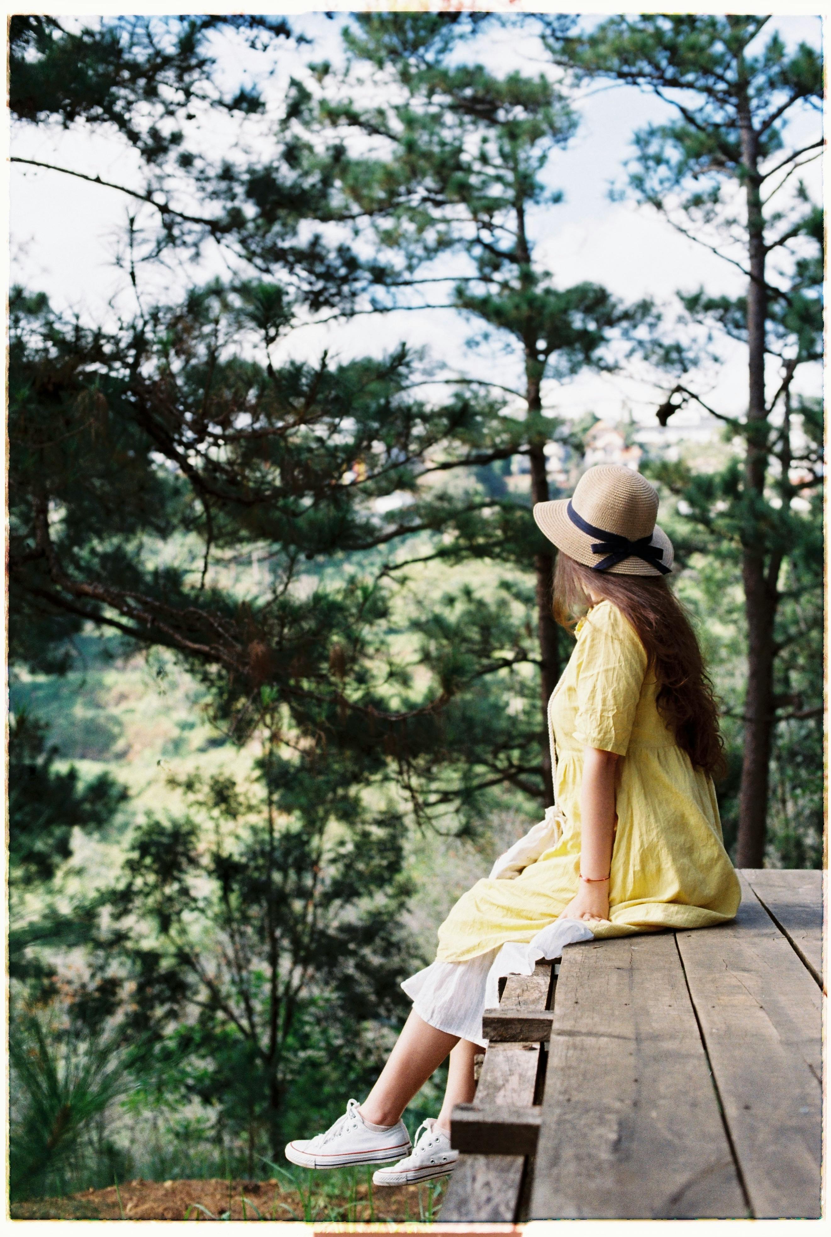 A woman in a yellow dress and hat sits on a wooden ledge, surrounded by trees.