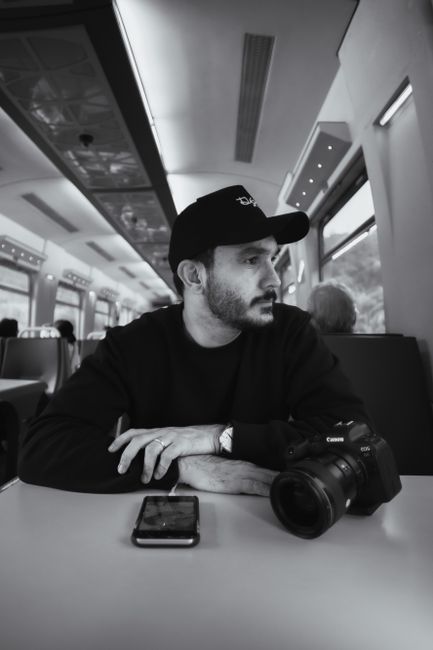 A man in a cap sits in a train with a camera and cellphone, captured in black and white.